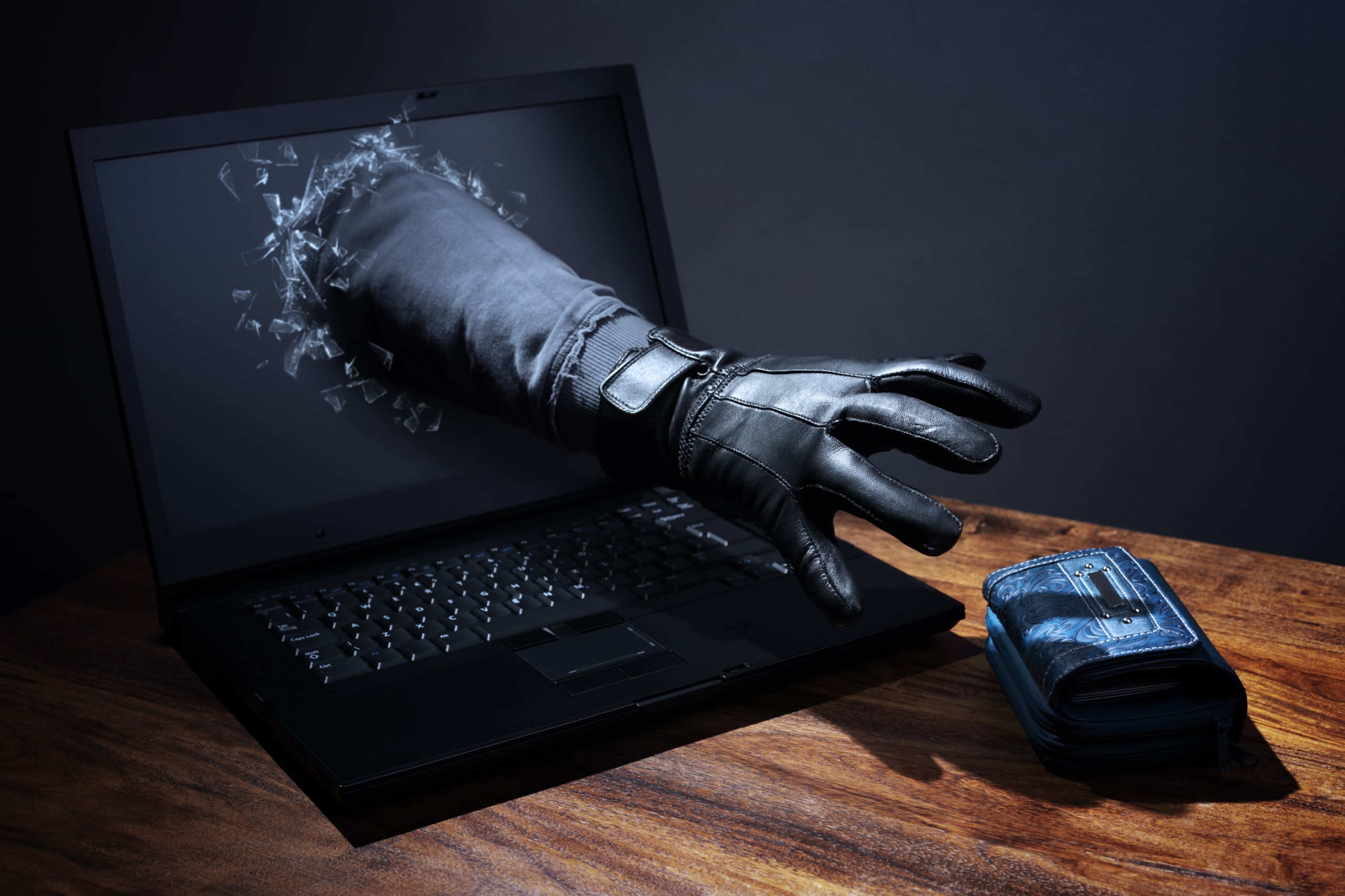 Dark gloved hand reaching through a laptop screen for a wallet that's placed on a desk next to the laptop